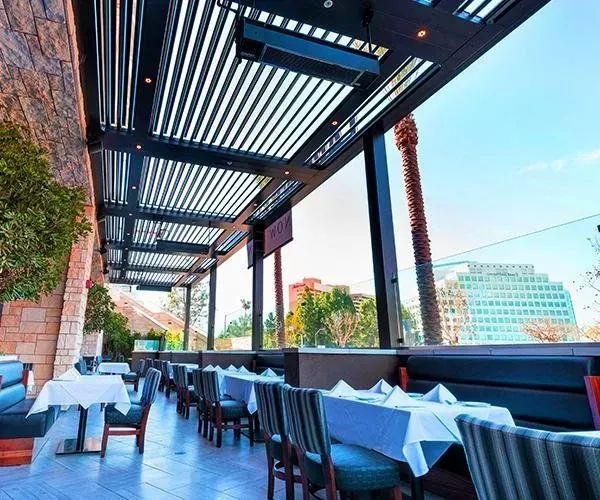 Outdoor restaurant seating under a modern, slatted roof. Tables are set with white linens. City view.