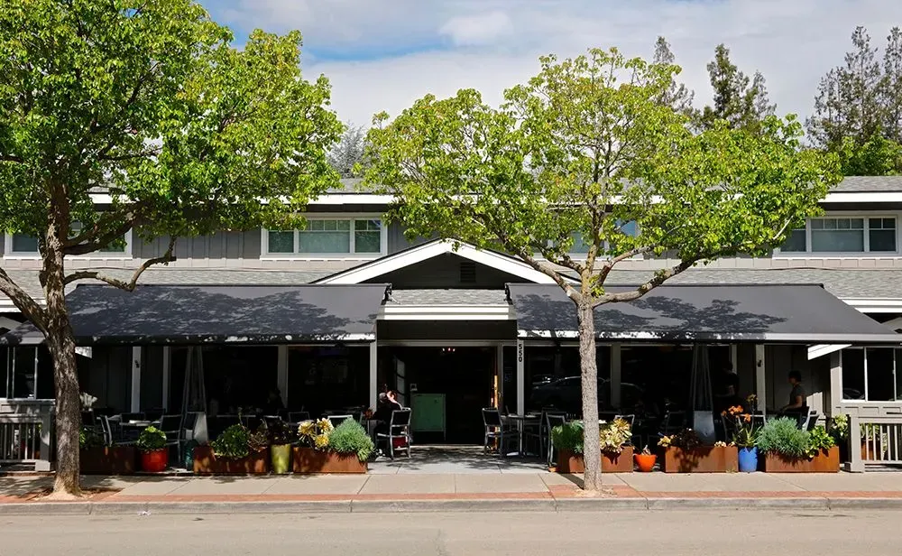 Restaurant exterior with black awnings, trees, and potted plants on the sidewalk.