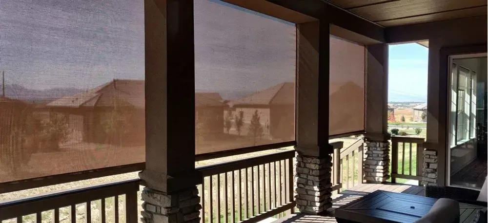 Covered patio with retractable shades overlooking a landscape. Brown wooden beams and stone columns frame the view.