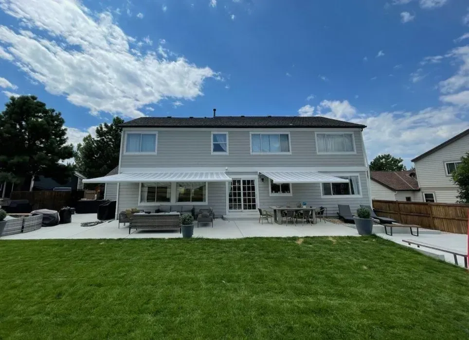 Backyard view of a two-story gray house with awnings, a patio with furniture, and a green lawn under a blue sky.