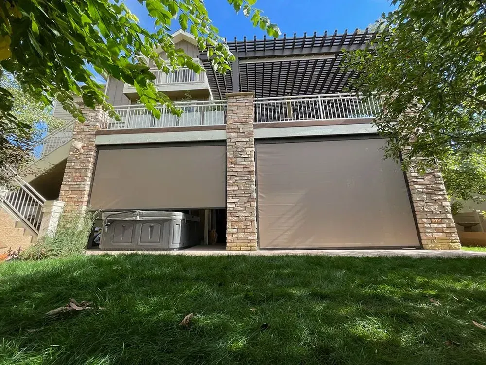 Tan retractable shades enclose a patio, with a hot tub visible below the upper deck. Green lawn and trees surround the house.