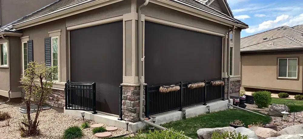 Beige house with a covered patio, dark brown shades, and a black wrought iron fence.