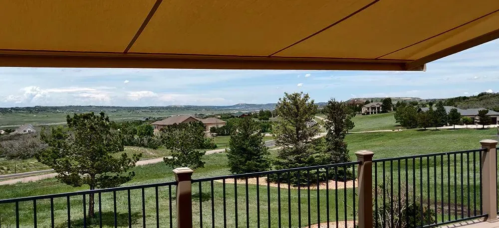 View of a grassy field, trees, and houses from a deck with a black railing, under a yellow awning.