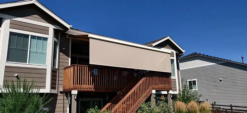 A wooden deck with a beige awning attached to a two-story brown house with a blue sky background.
