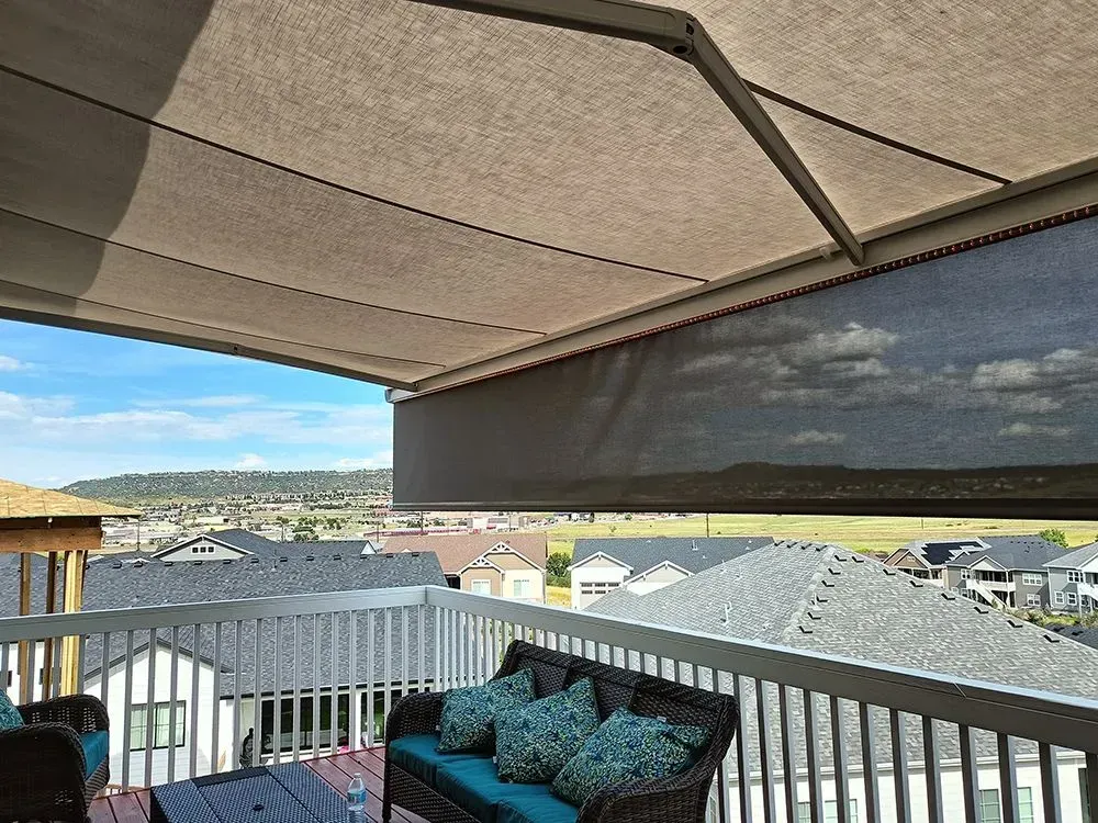 A beige retractable awning covers a deck with a view of houses and a blue sky.