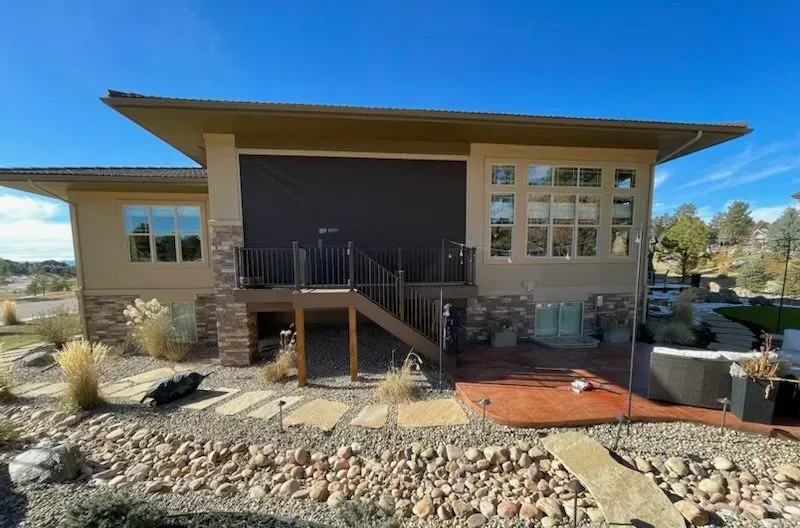 Rear view of a beige house with a brown deck, black sun shade, and hot tub on a sunny day.