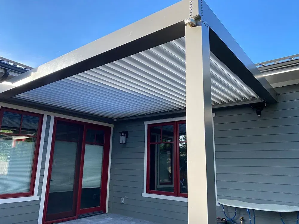 Gray aluminum pergola over a patio. Gray siding, red-framed windows, blue sky.