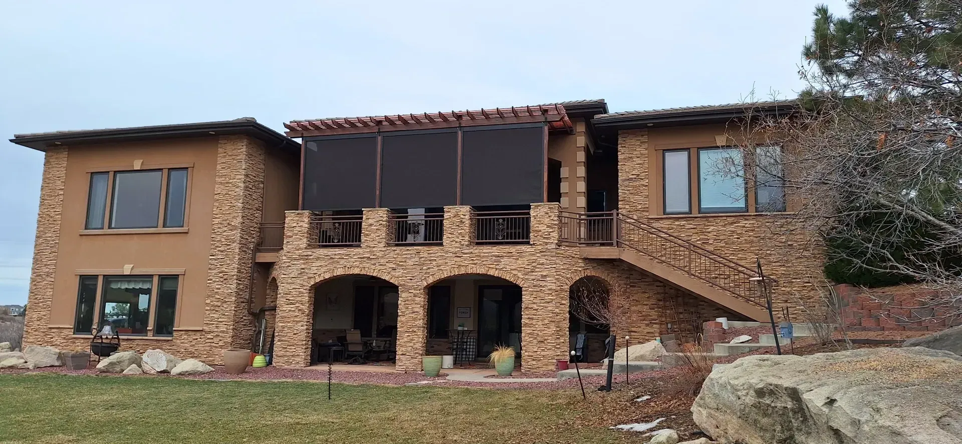 Two-story house with stone exterior, arched entry, and dark sun screens, under an overcast sky.
