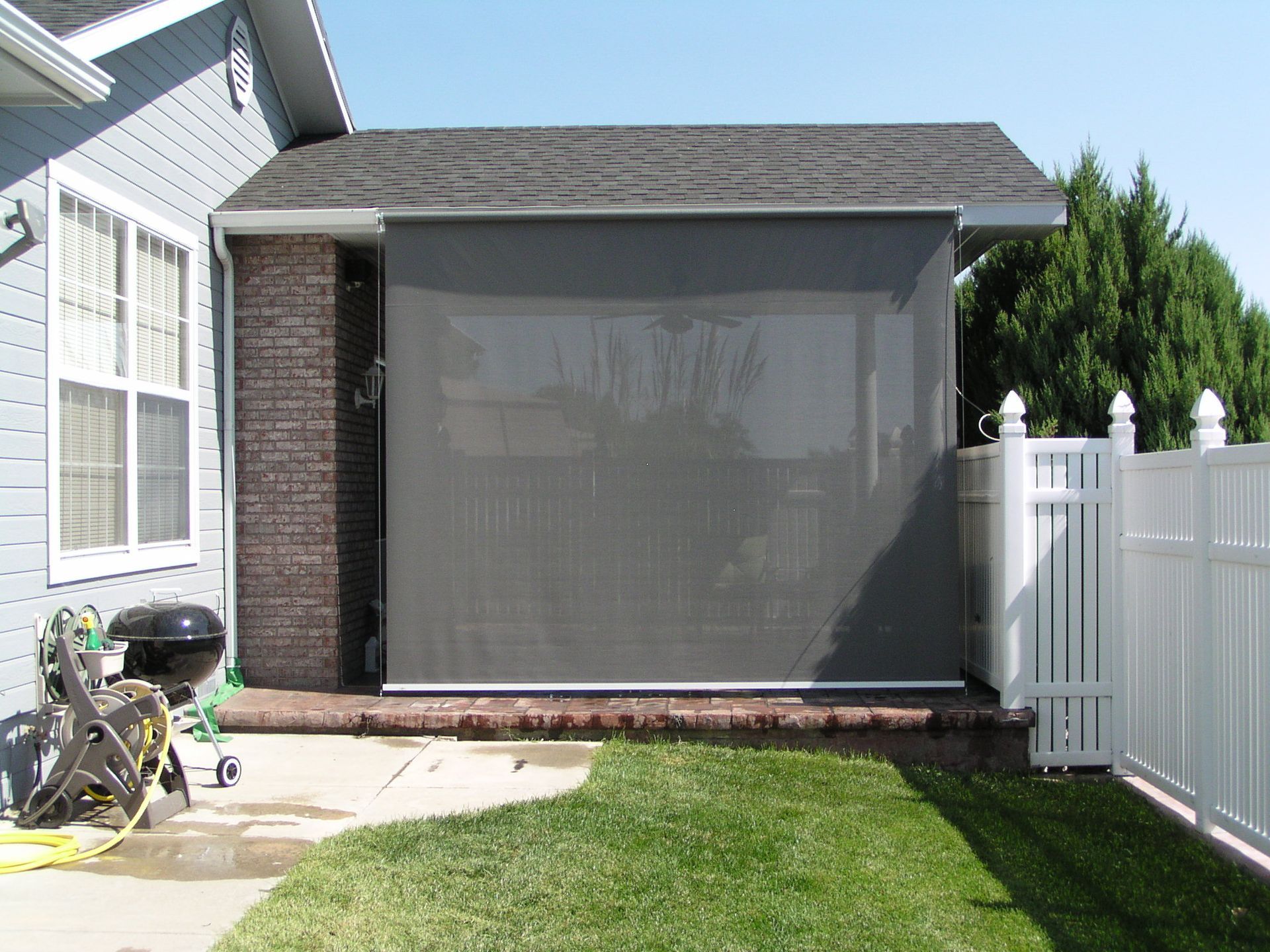 Dark gray outdoor sun shade attached to a brick wall and a roof, beside a white fence.