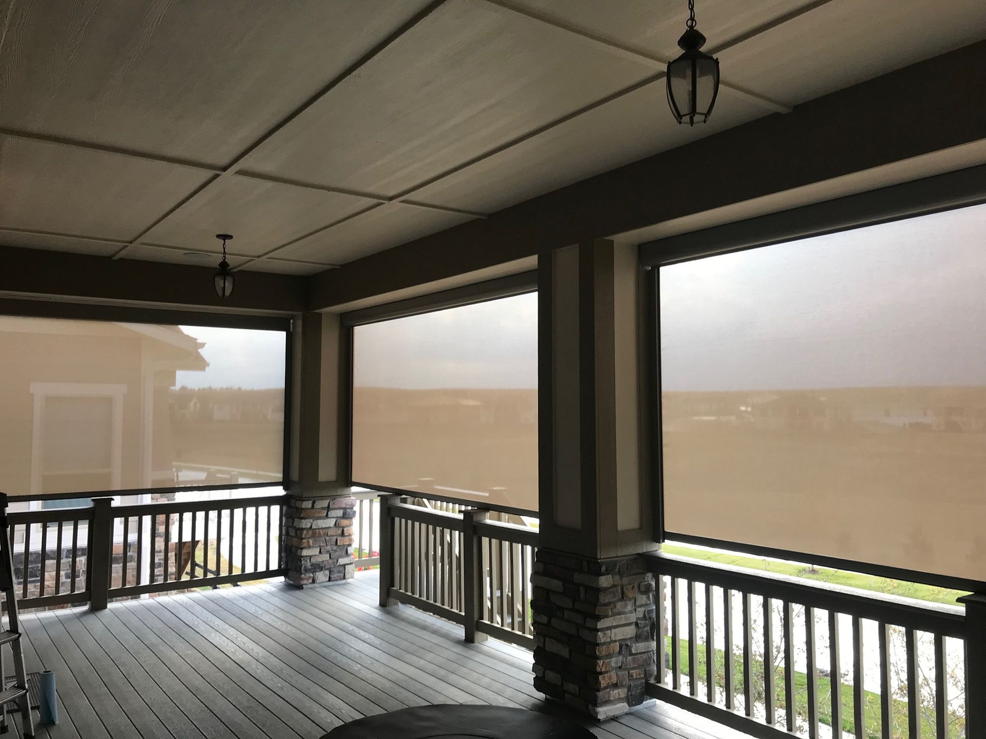 Covered patio with roll-down shades. Beige shades block sunlight. Wooden deck and railings.
