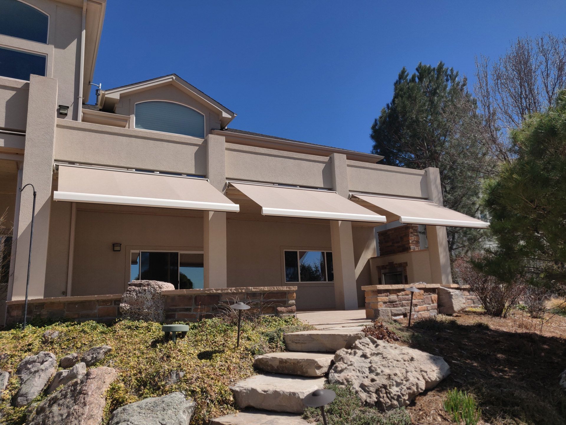 Beige house with three retractable awnings over windows, stone steps, blue sky.