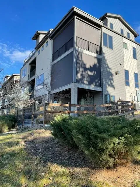 Three-story apartment building with gray siding and balconies beside a grassy slope under a blue sky