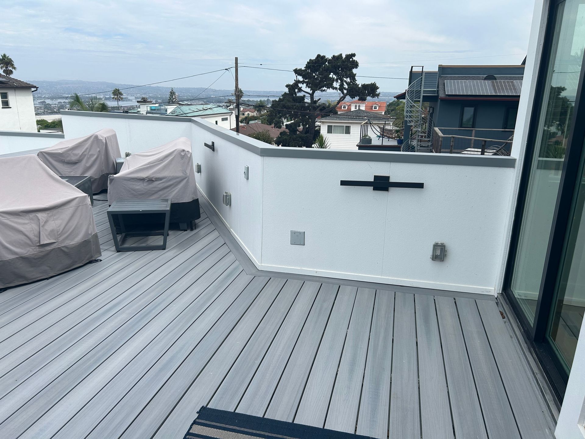 Rooftop deck with gray planks, white walls, and covered furniture; city view in the background.