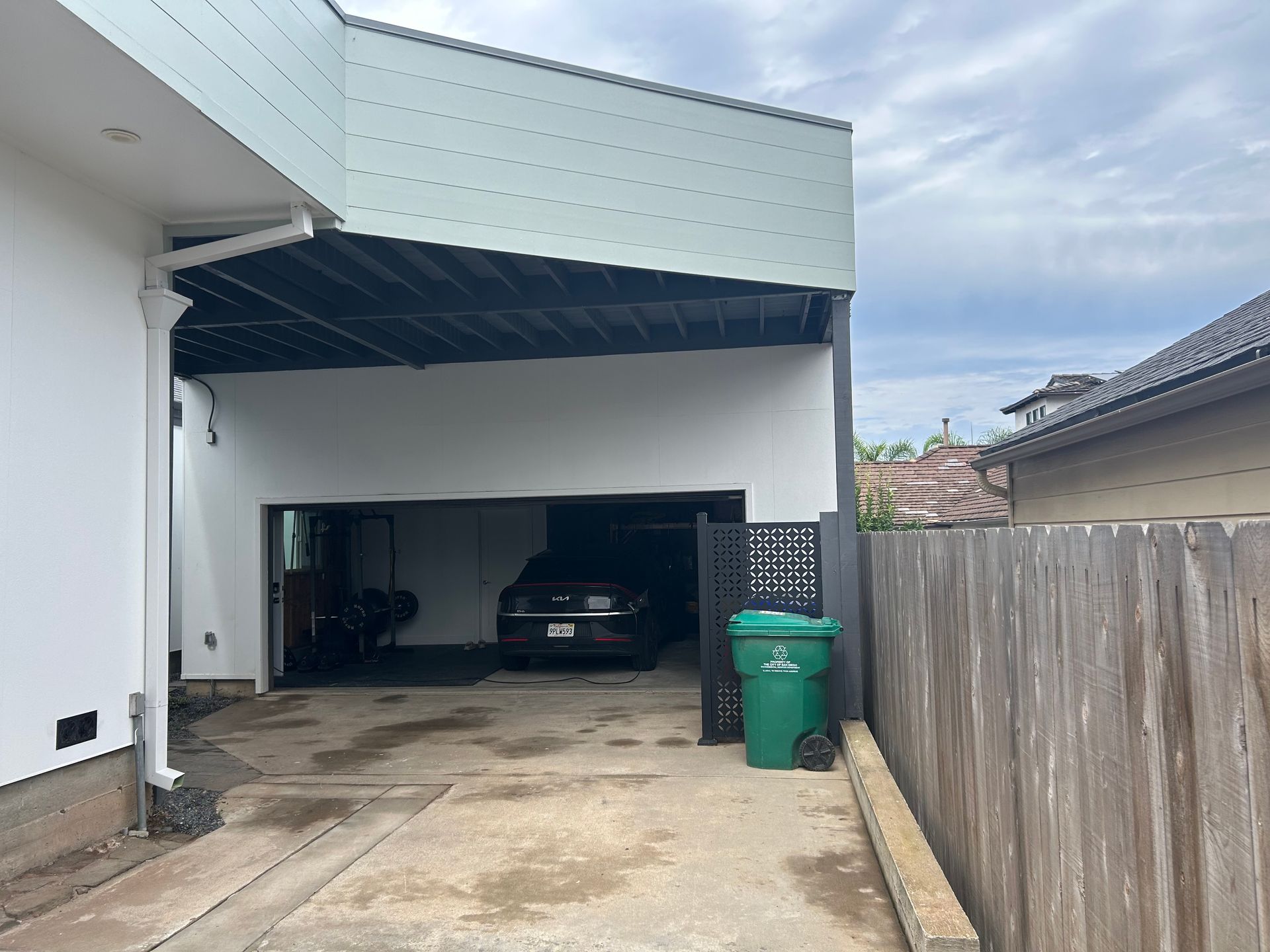 A garage with a car inside, next to a wooden fence. A green trash can sits near the fence.