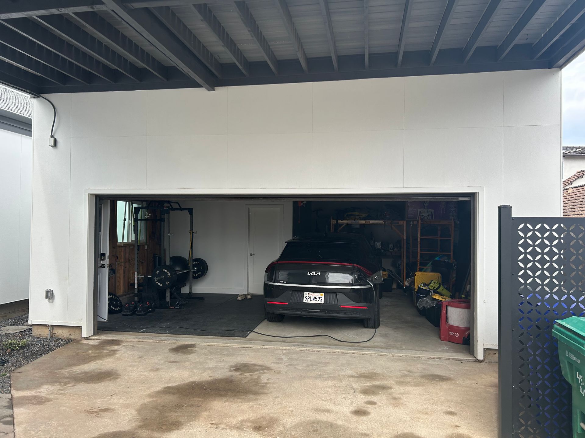 Garage exterior with a car inside and weights, a trash can, and decorative fence.