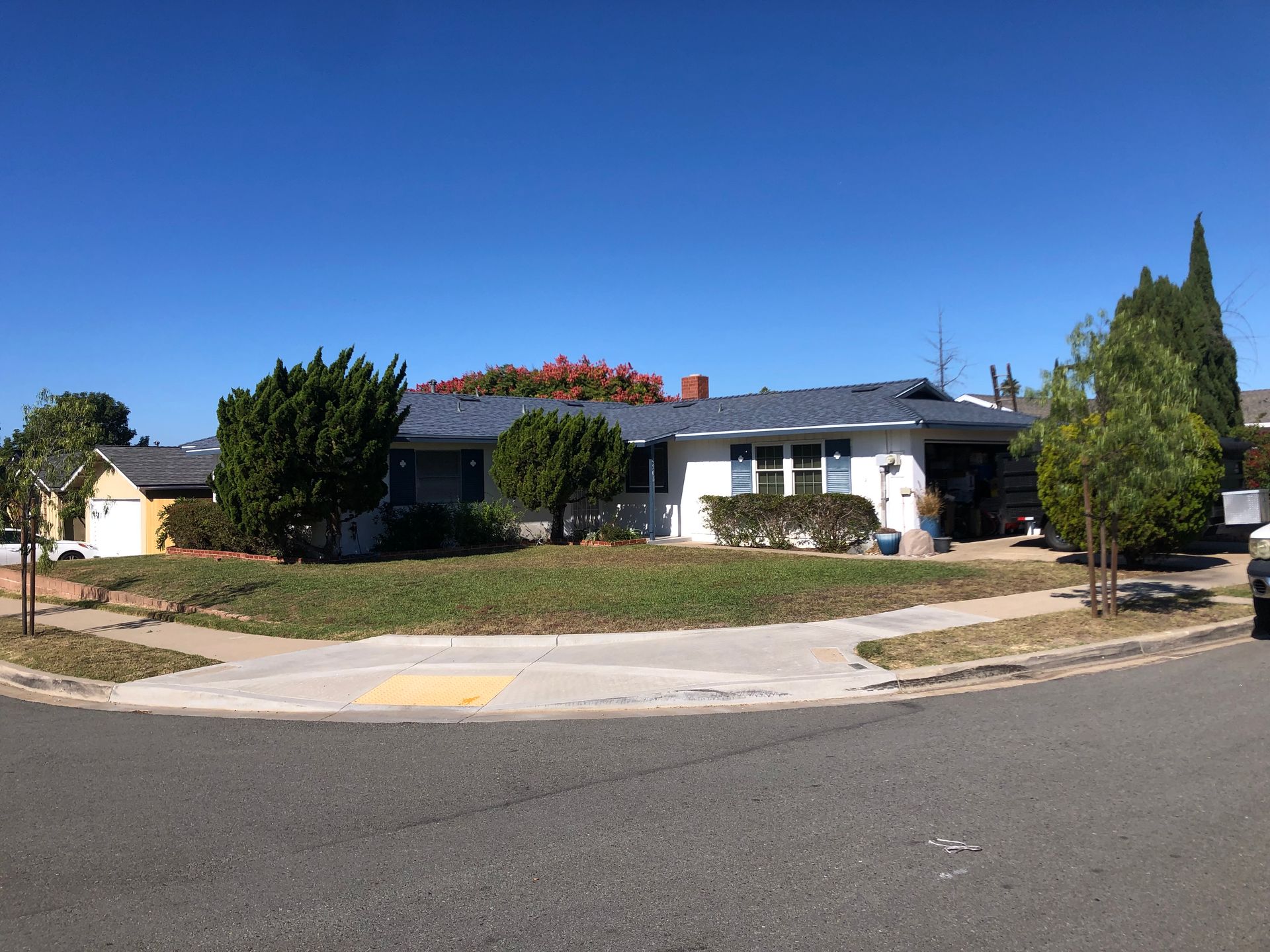 White house with blue trim on a sunny day, facing the street. Trees and shrubs in front yard.