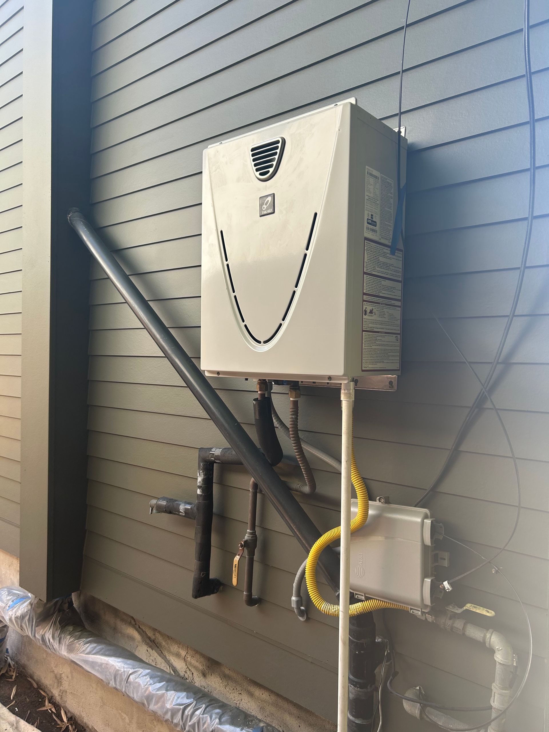 Tankless water heater mounted on a gray house exterior. Black pipes and yellow electrical lines connect to it.