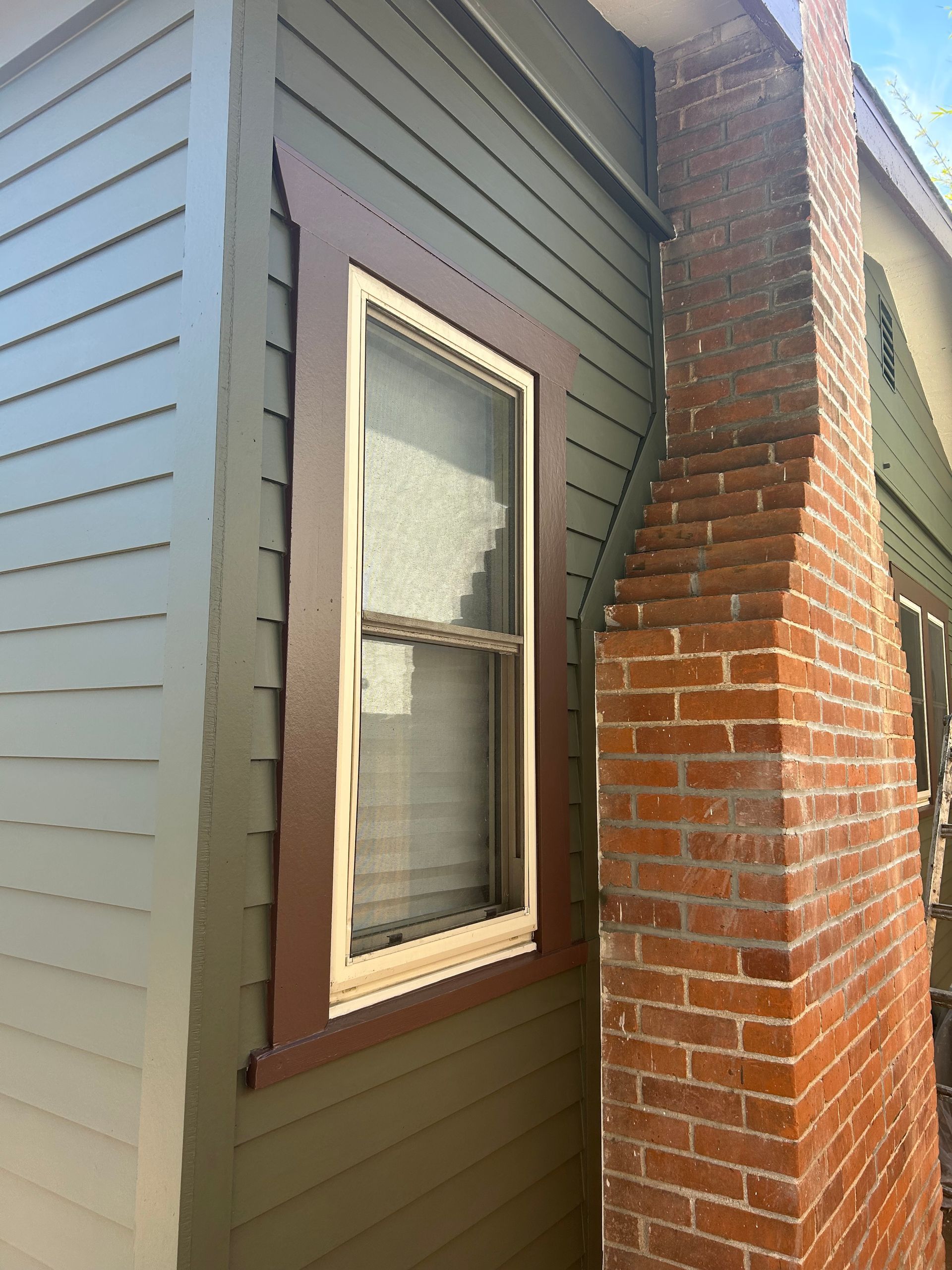 Green siding with brown trim around a window and a brick chimney.