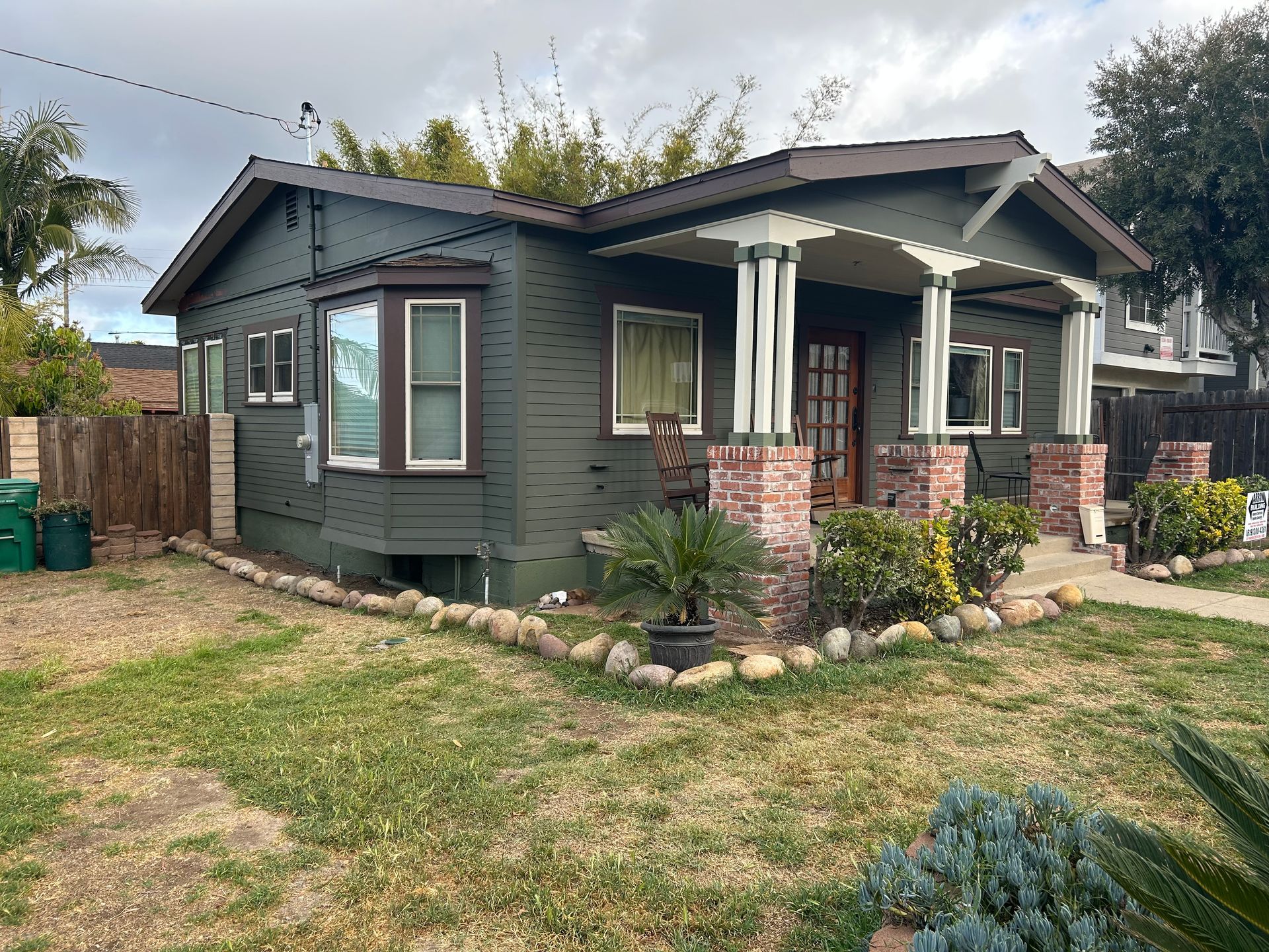 Green Craftsman house with white trim, bay window, and brick porch on a grassy lot.