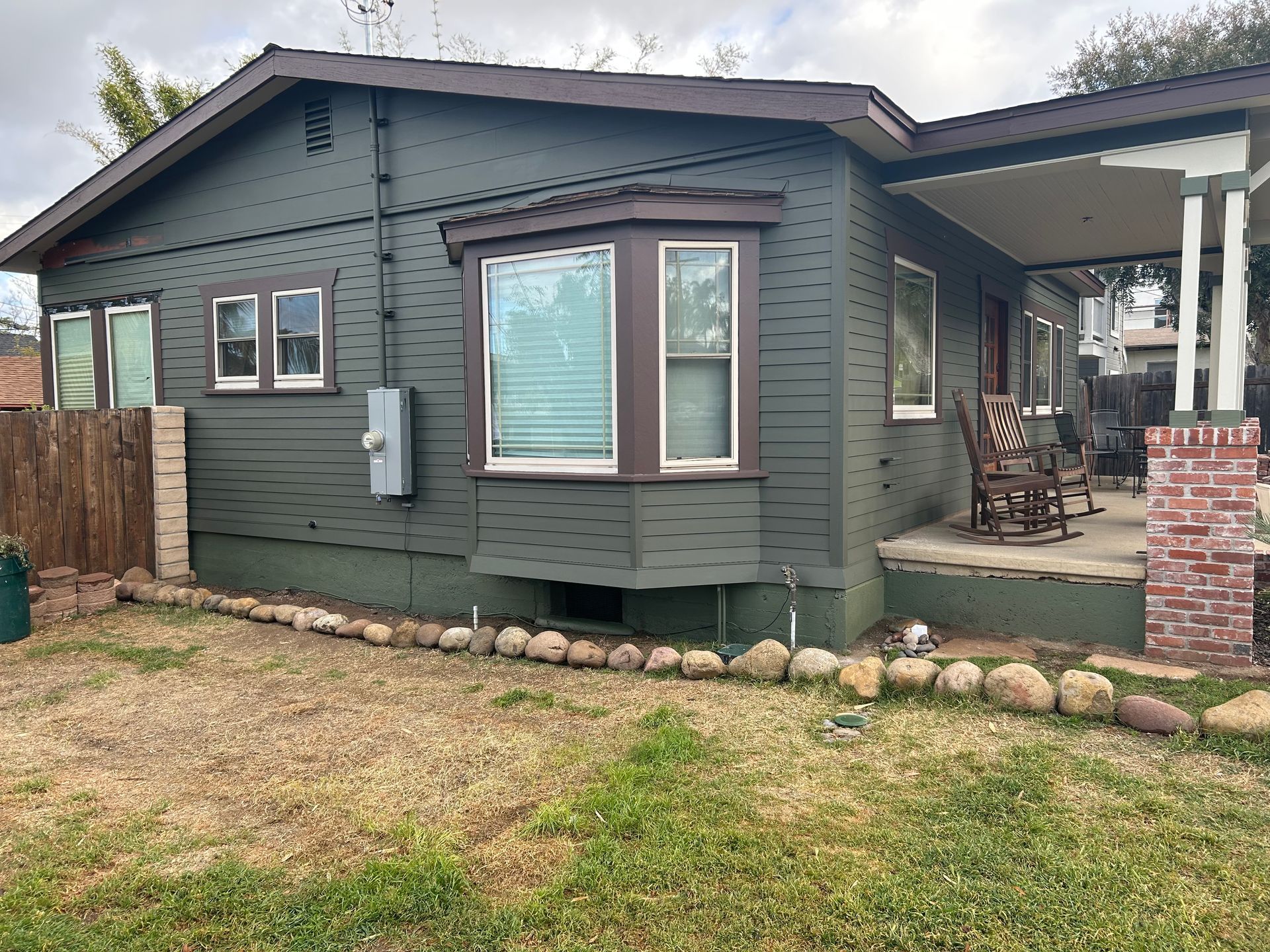 Green house with porch and bay window, brown trim, in front of a yard with rocks.