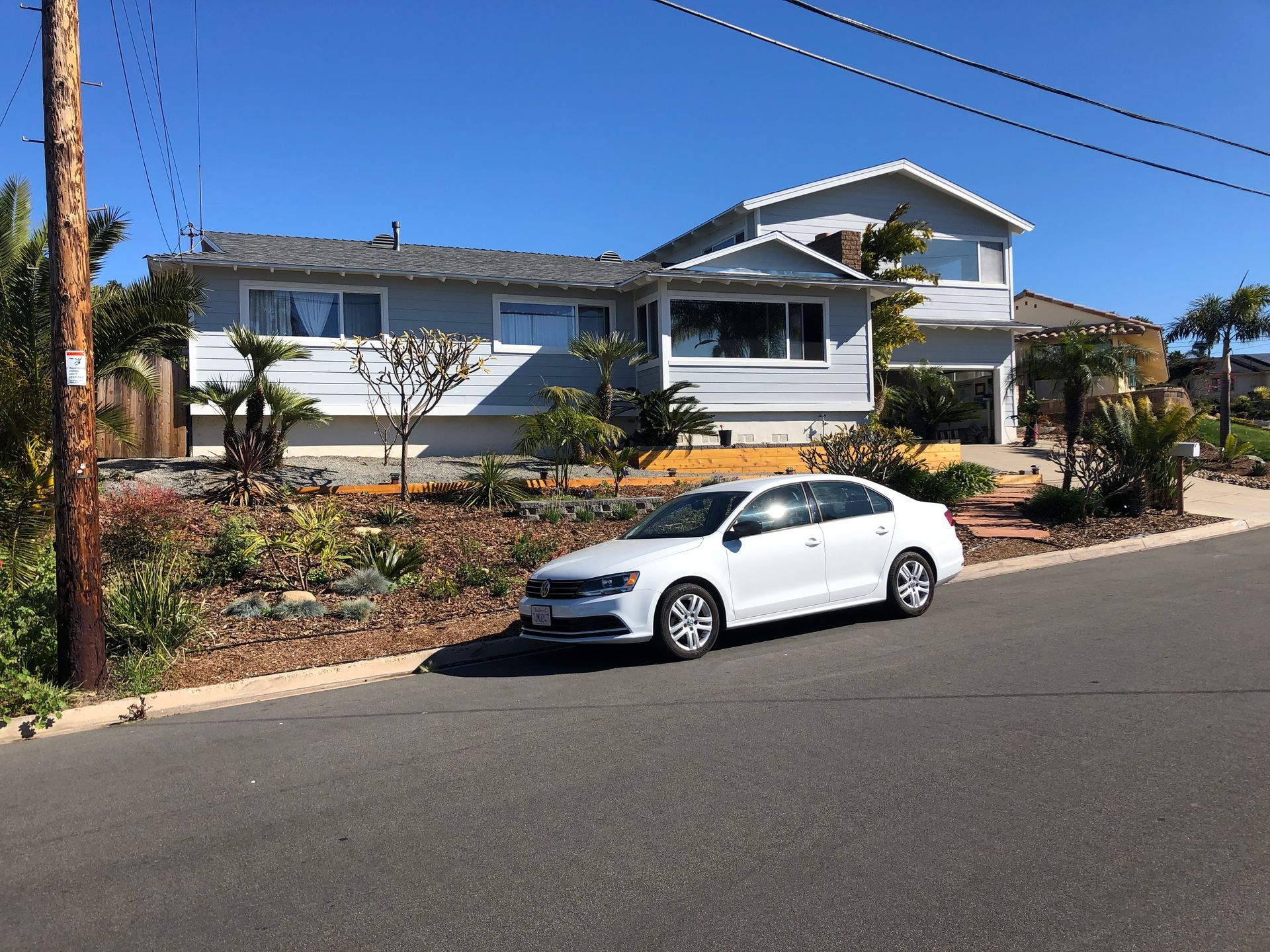 White car parked on a sloped street in front of a light blue house with a two-story addition on a sunny day.