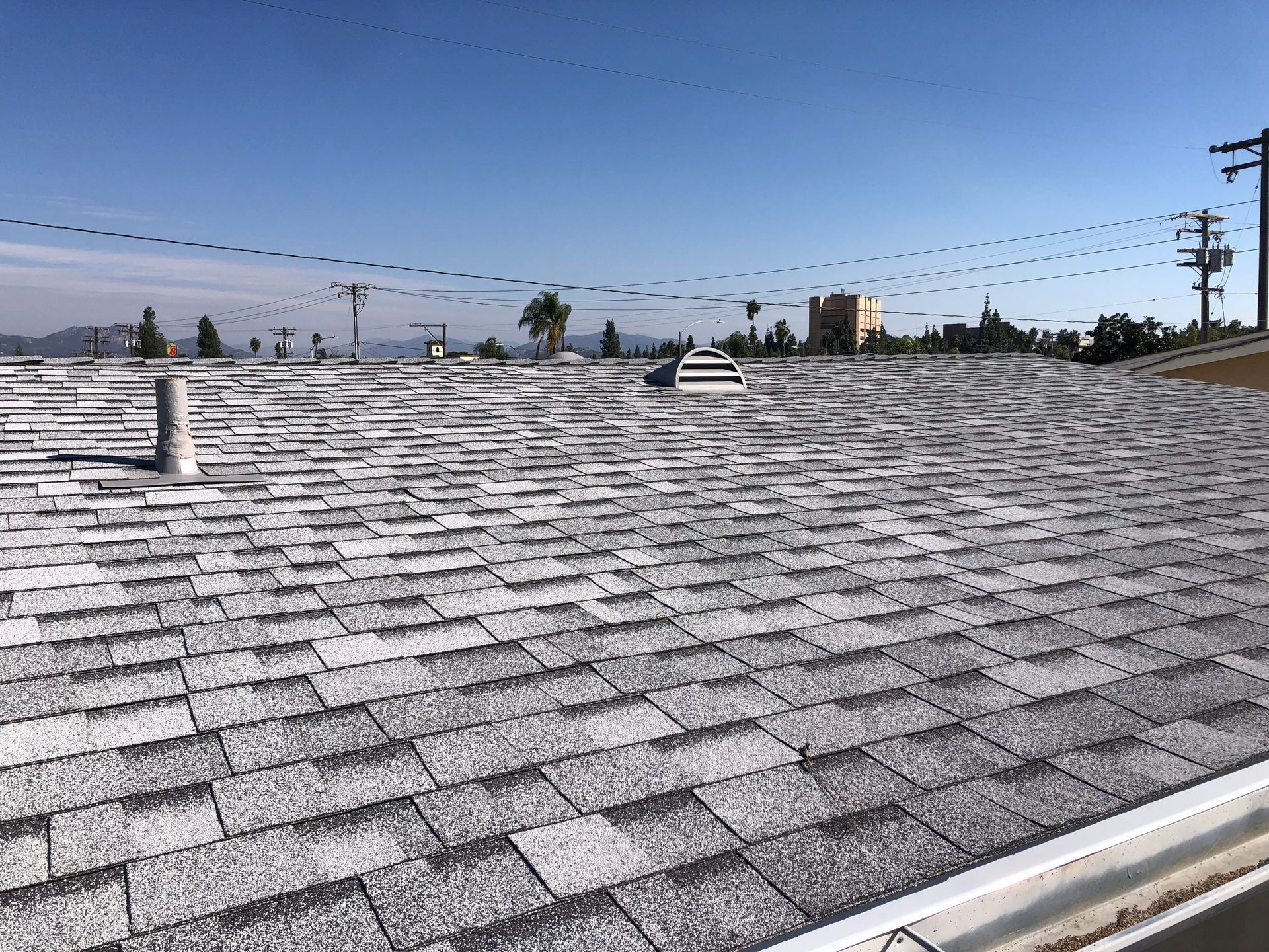 A grey asphalt shingle roof on a house, clear blue sky in the background, power lines visible.