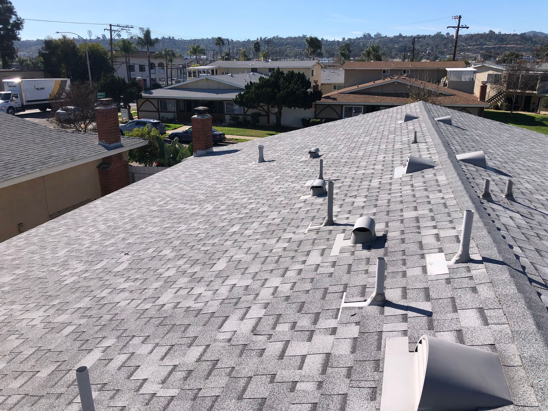 View of a light gray shingle roof with pipes, chimneys, and a suburban background on a sunny day.
