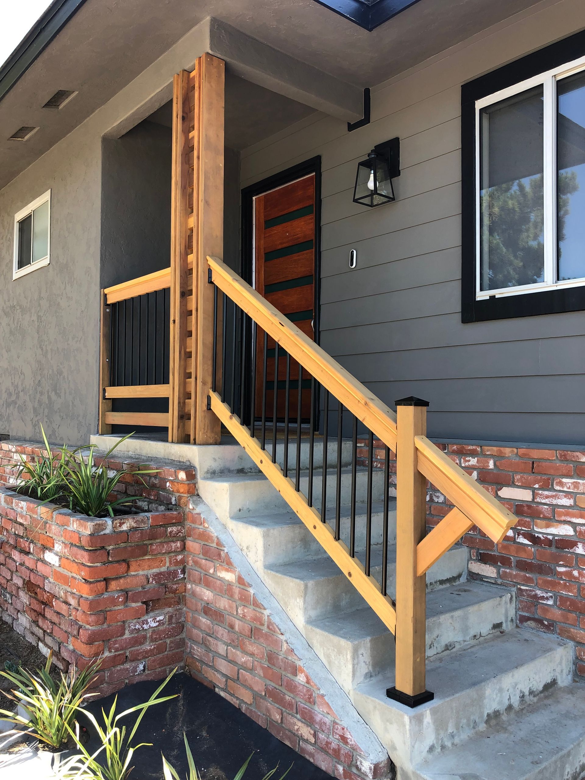Entryway with concrete steps, brick planter, wooden railing, brown door, gray siding.