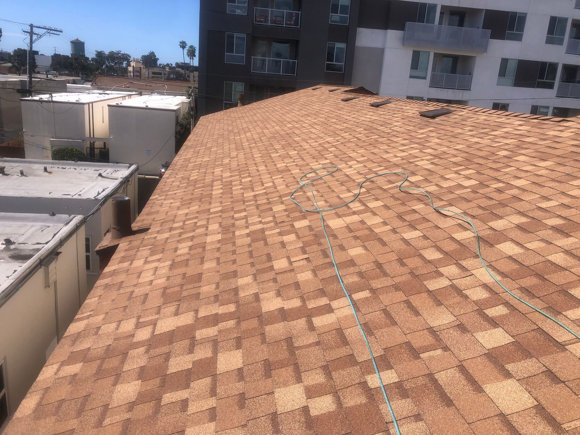 Brown shingle roof with a multi-story apartment building in the background, sunny day.