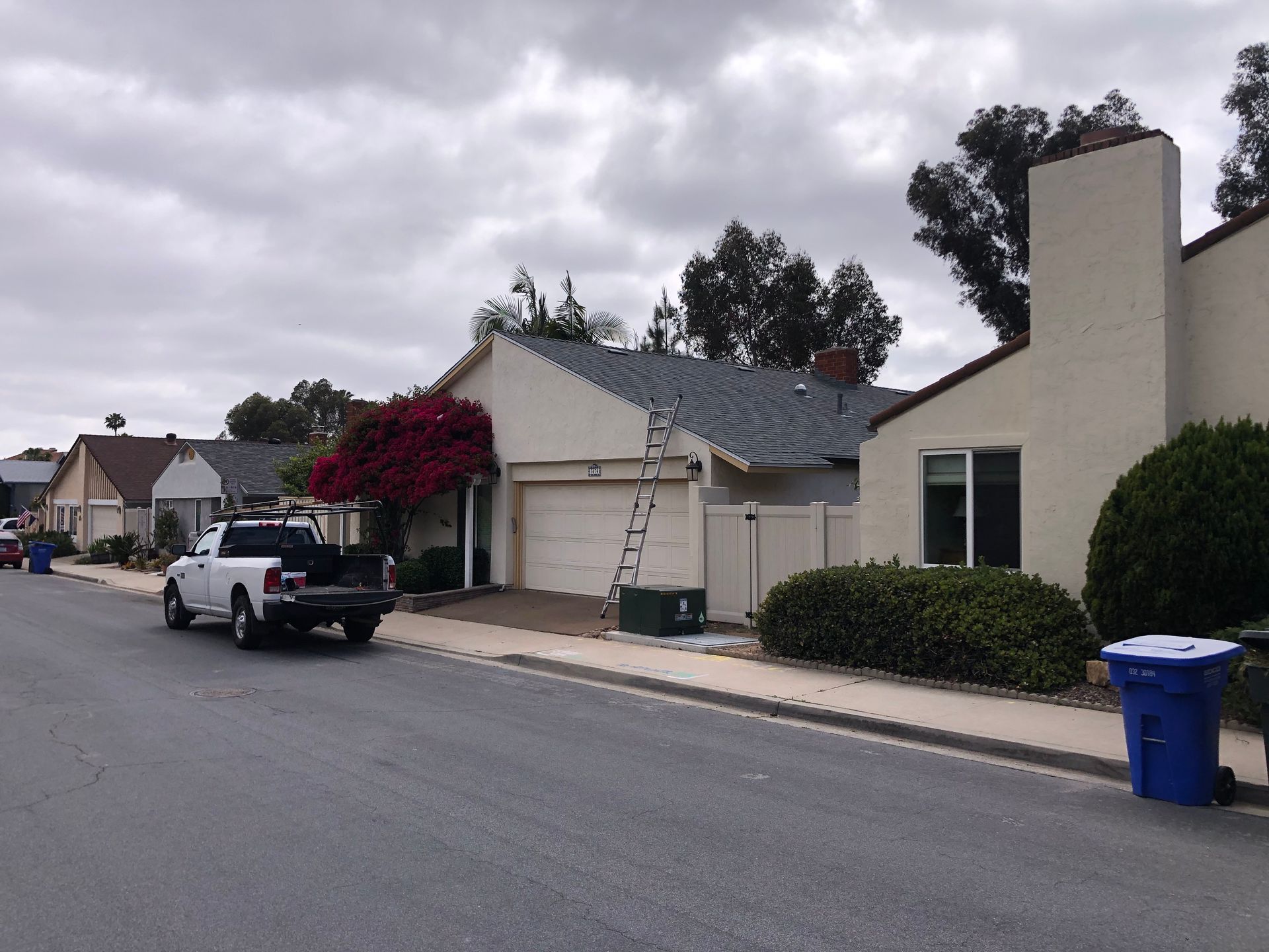 Street view of houses with a white truck, ladder, and trash bins on a cloudy day.