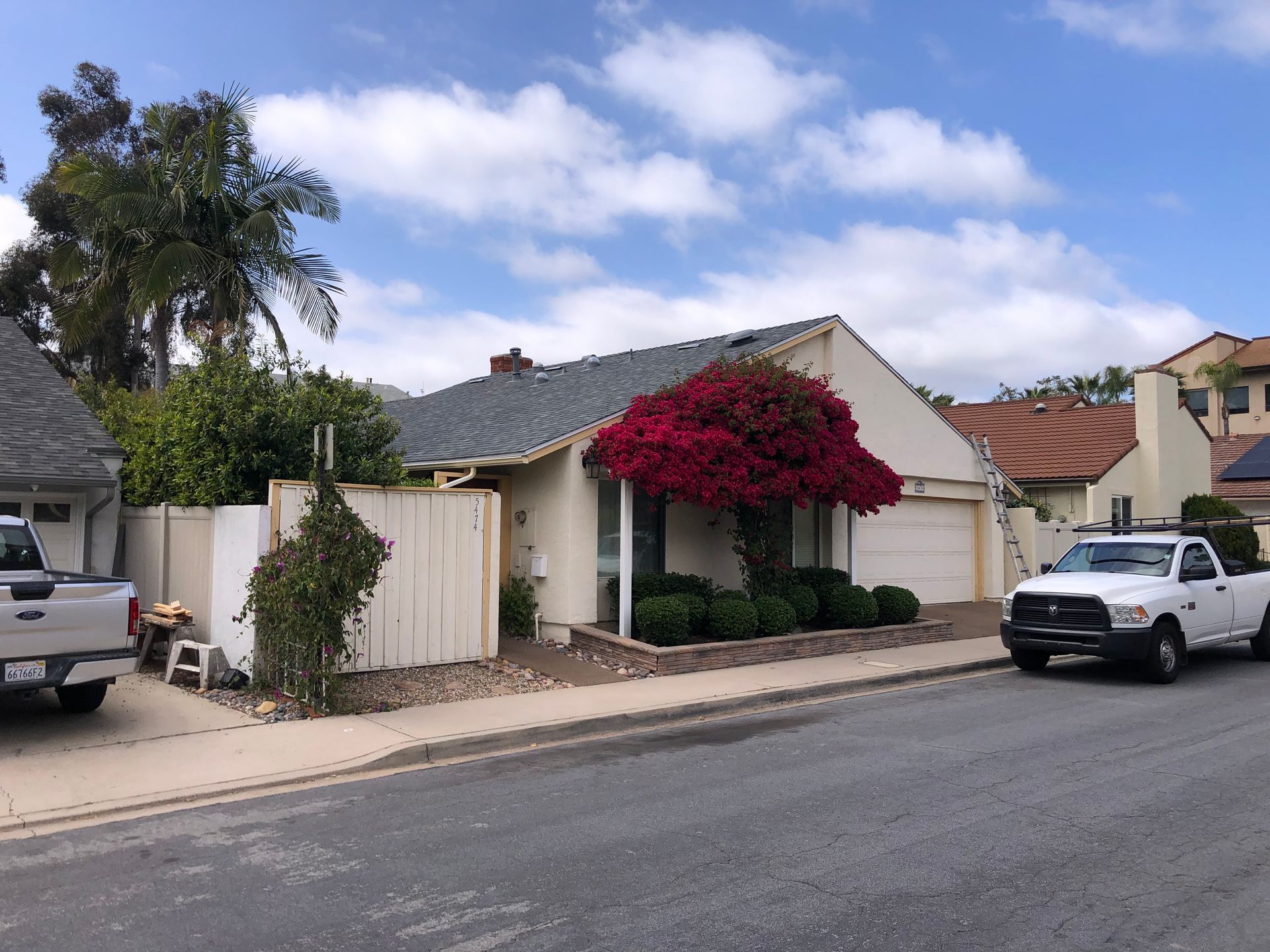 Residential street scene: beige house with red bougainvillea, white truck, blue sky.
