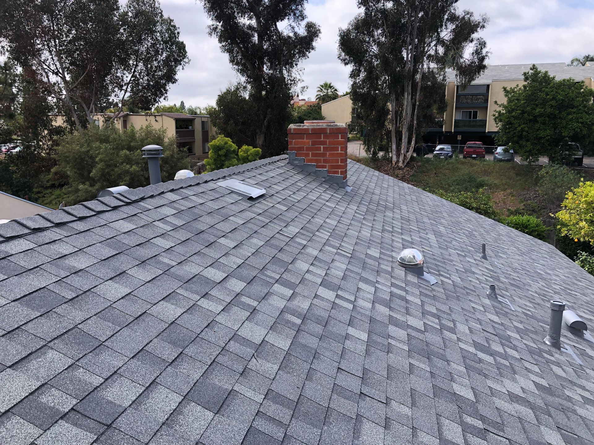 A gray asphalt shingle roof with a red brick chimney, trees, and overcast sky.