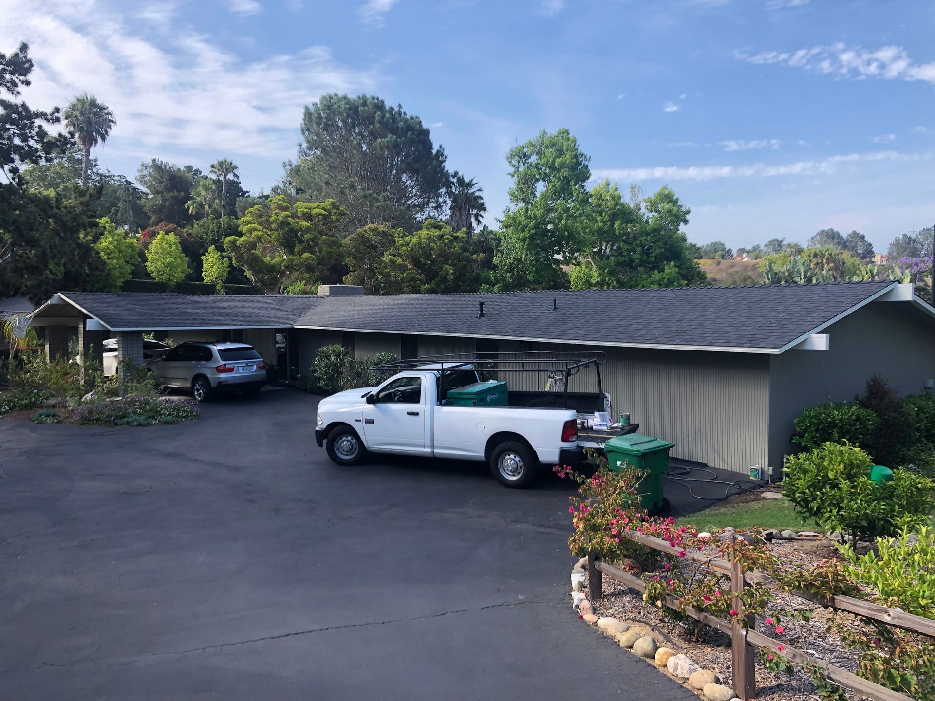 A house with a black roof, driveway with a car and truck parked. Green foliage in the foreground.