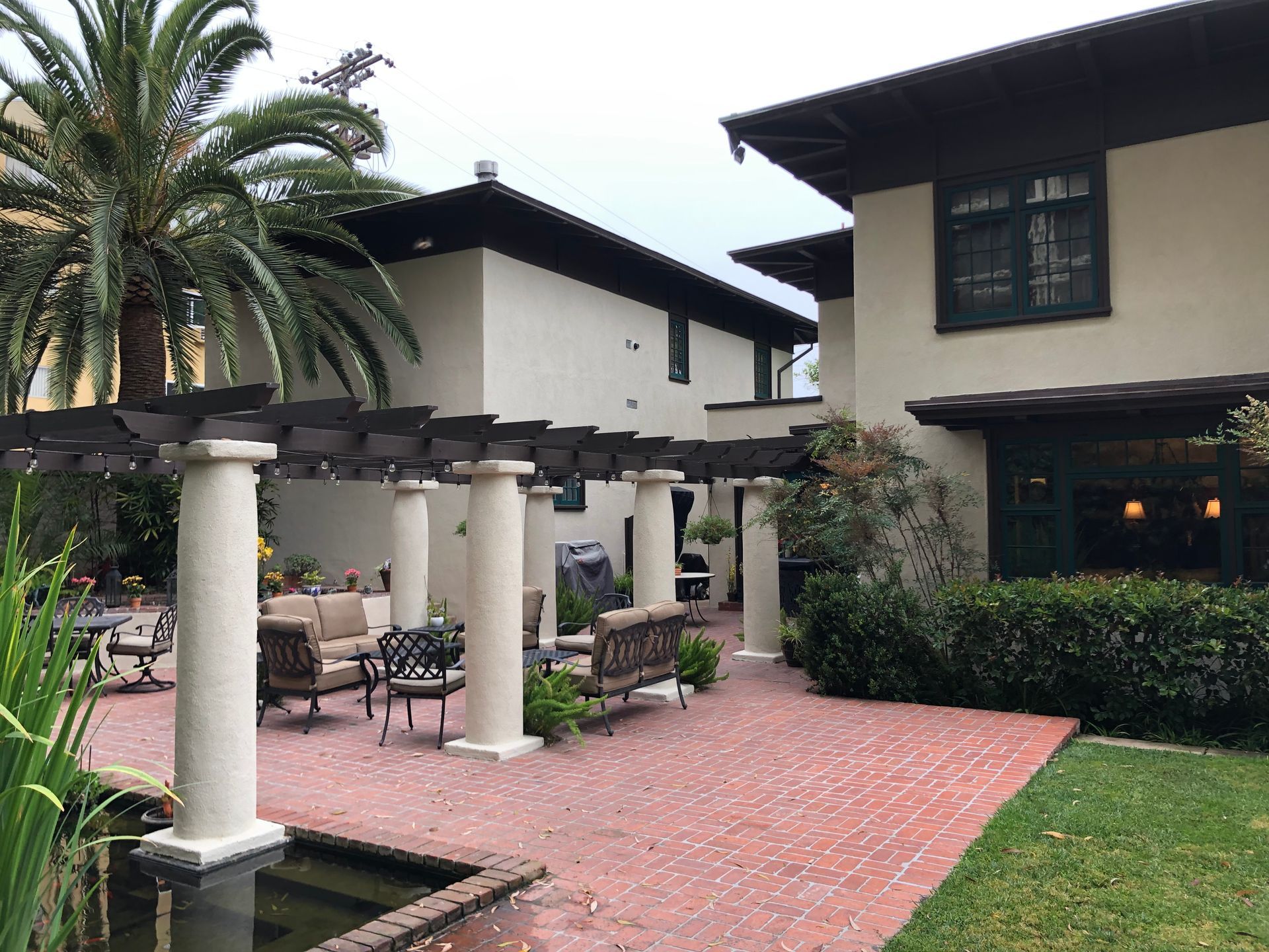 Courtyard with pergola, seating, and buildings. Red brick patio, green lawn, and palm tree.