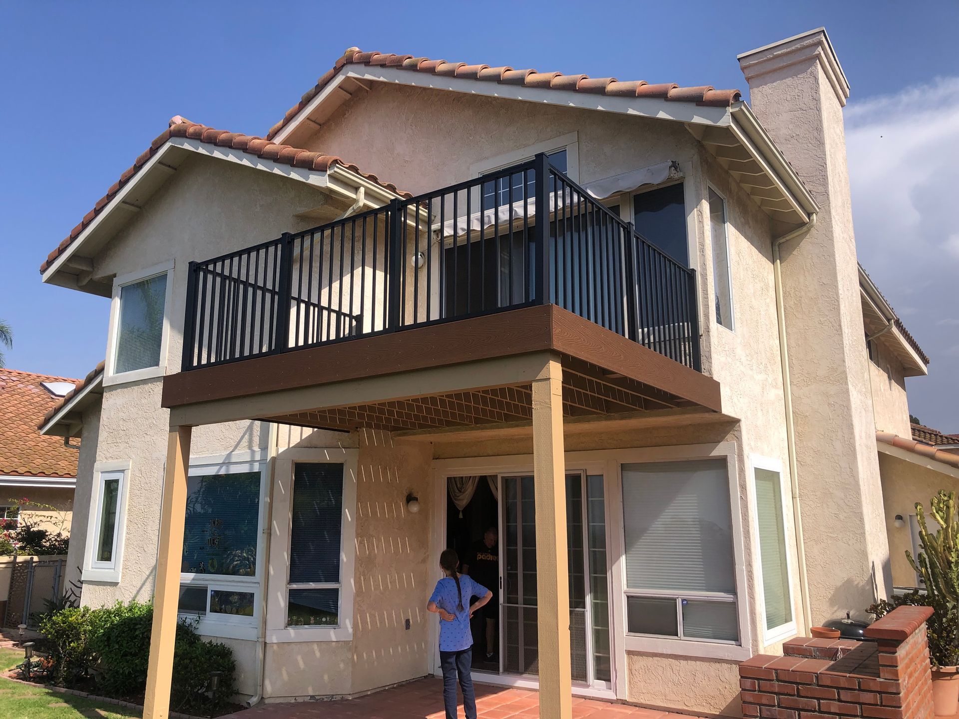 A two-story house with a deck. Person stands on patio. Brown deck, black railing, blue sky.
