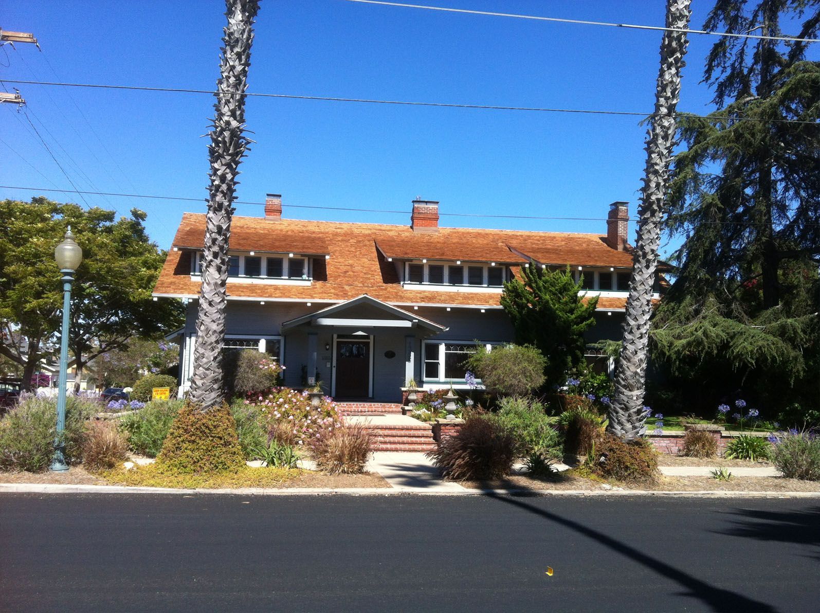 Gray house with brown roof, surrounded by trees and shrubs, under a blue sky.