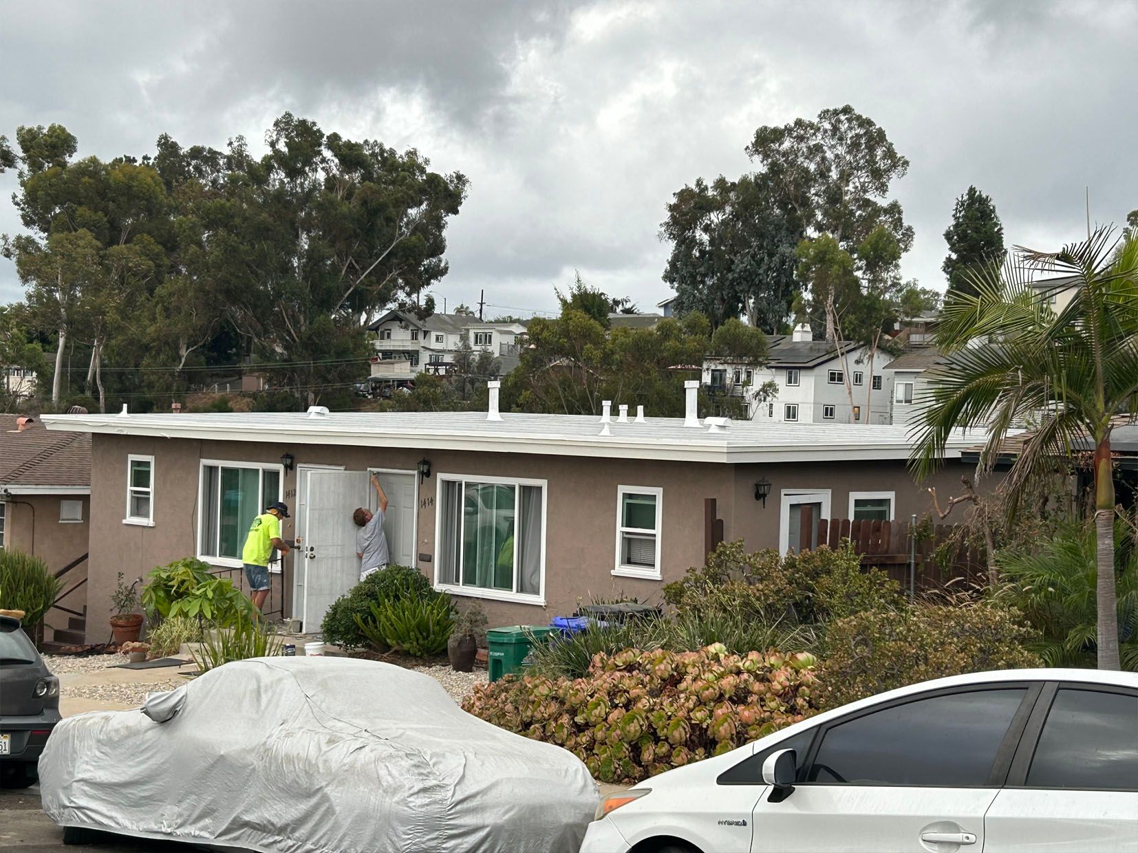 Two people at a brown, single-story house. One opens a door; a car is parked in front of the home.