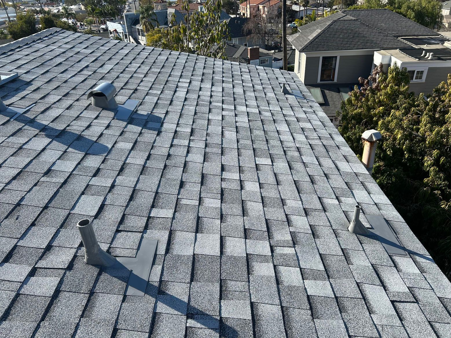 Gray asphalt shingle roof with vents, a sunny rooftop view of urban homes.