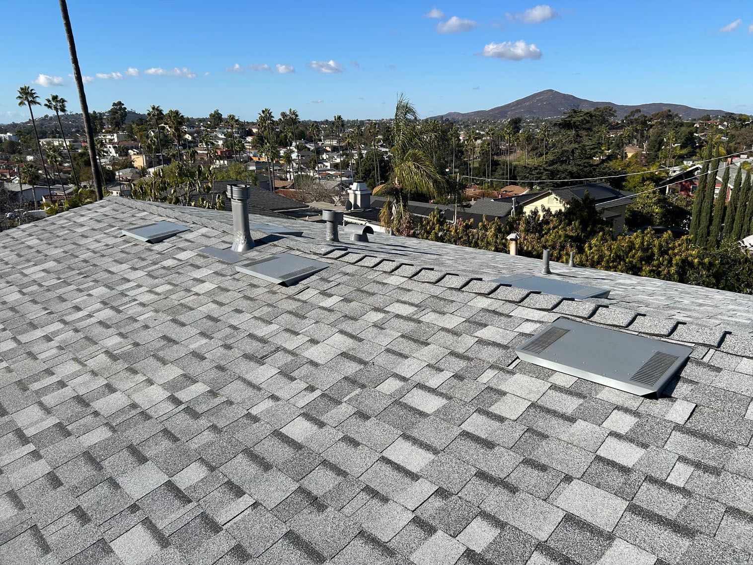 Gray shingle roof with vents and a cityscape view in the background under a blue sky.
