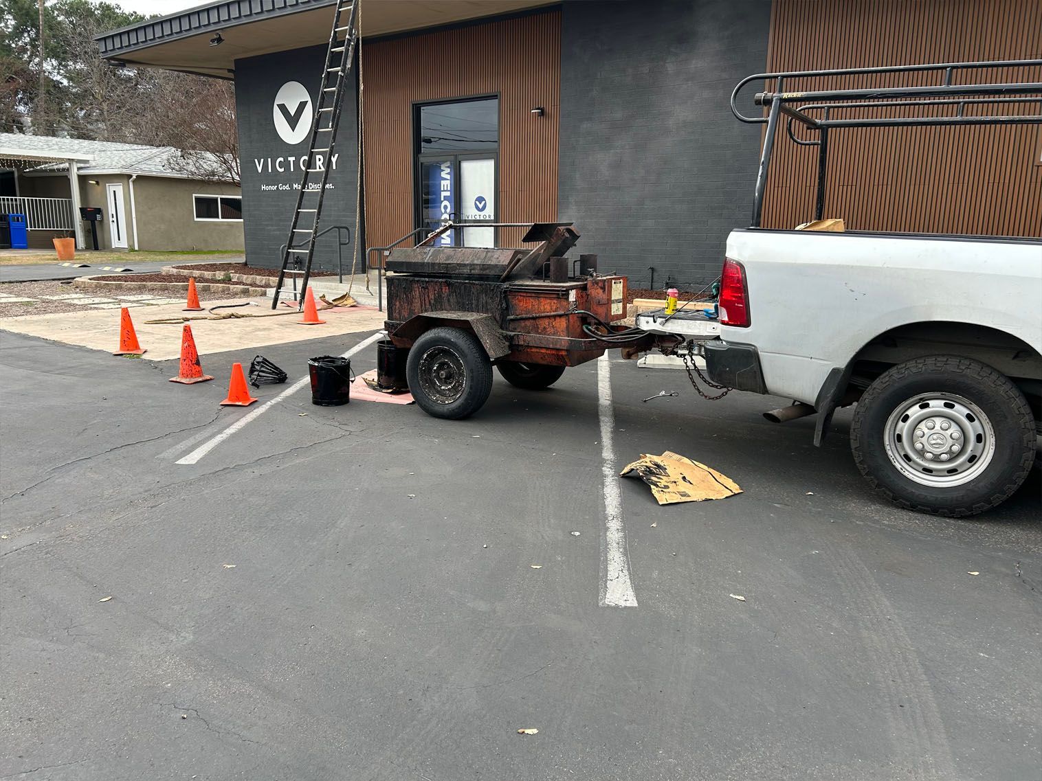White truck towing a rusty trailer parked outside a building. Construction cones mark the area.