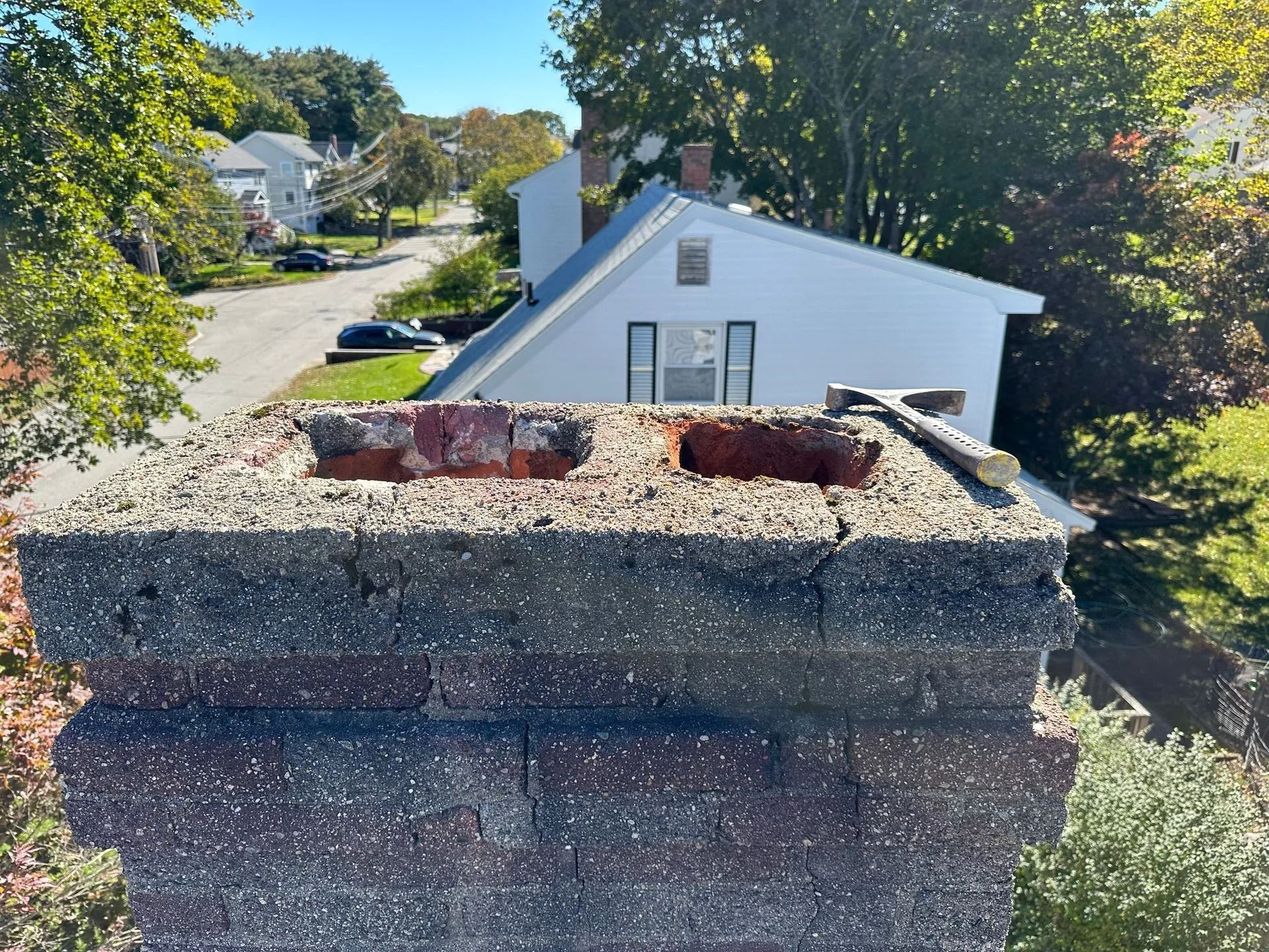 Chimney with damaged top, sitting on a roof. A hammer rests on the chimney. Houses and trees are in the background.