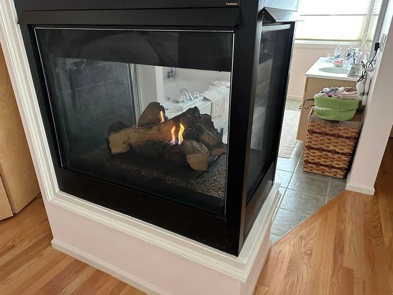 Gas fireplace with faux logs, in a corner, flames visible, adjacent to a bathroom.