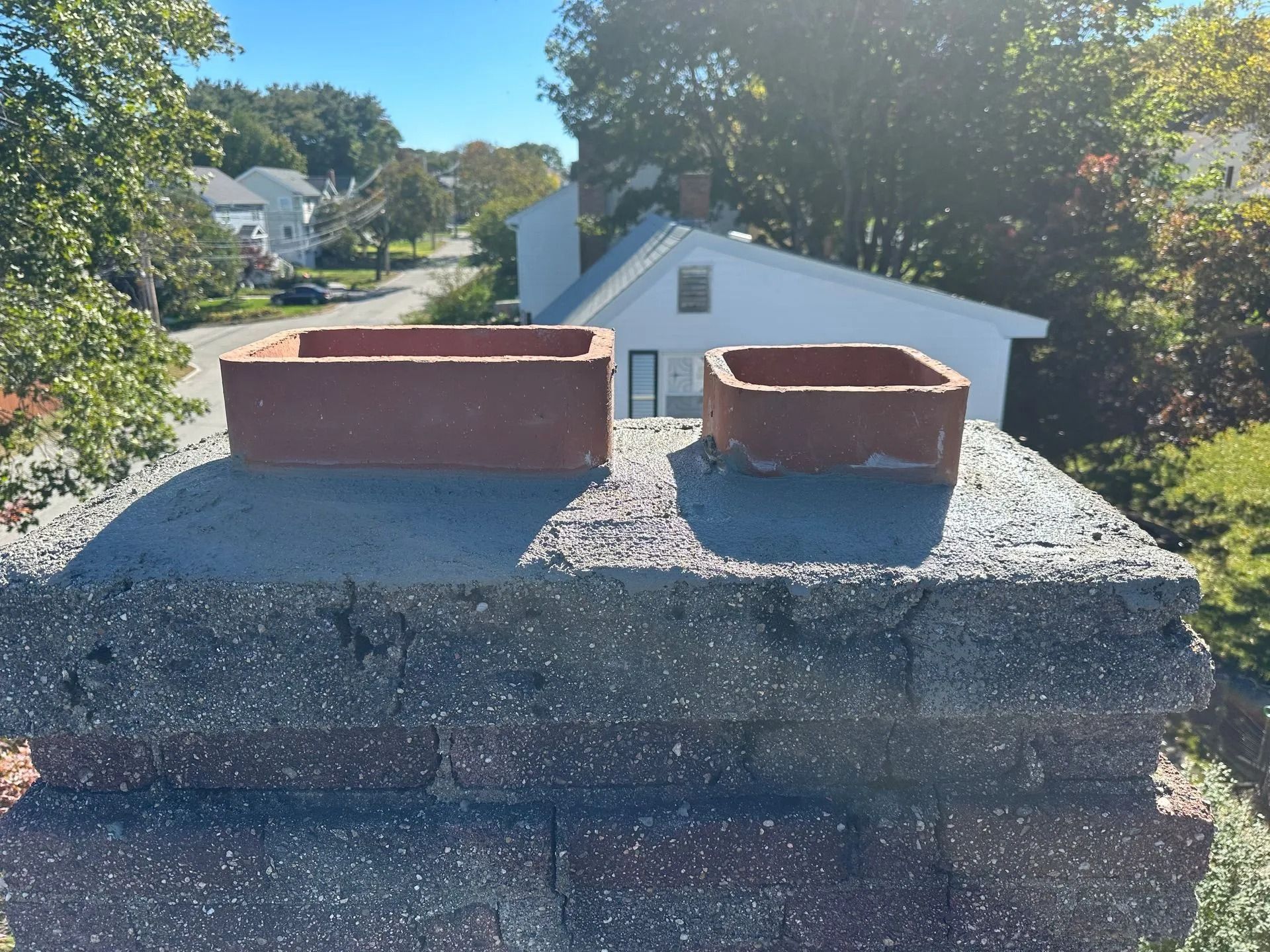 Two brick chimney caps on a brick chimney, with a tree and houses in the background on a sunny day.