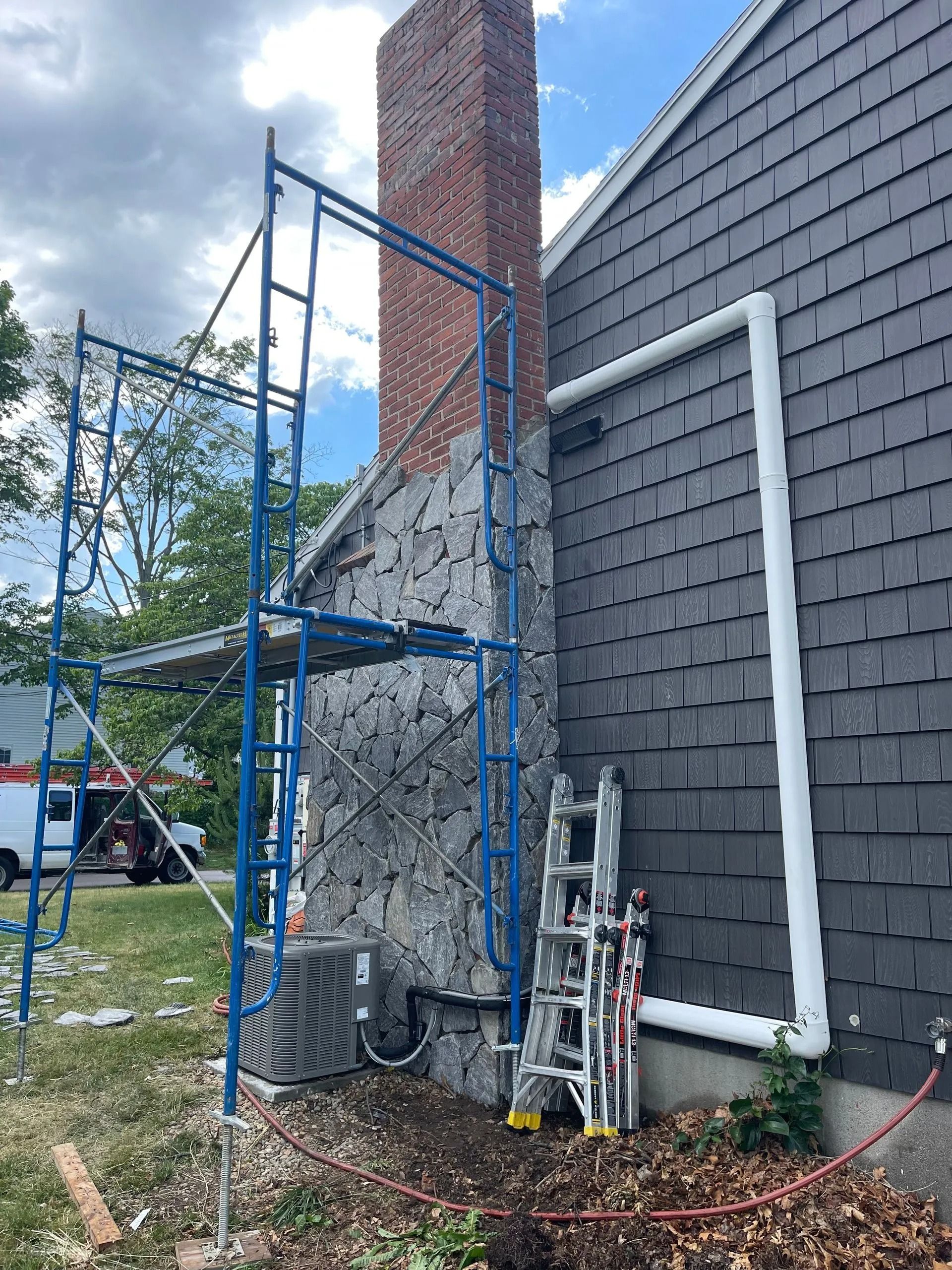 Scaffolding next to a brick chimney and house with gray shingles. A/C unit on the wall.