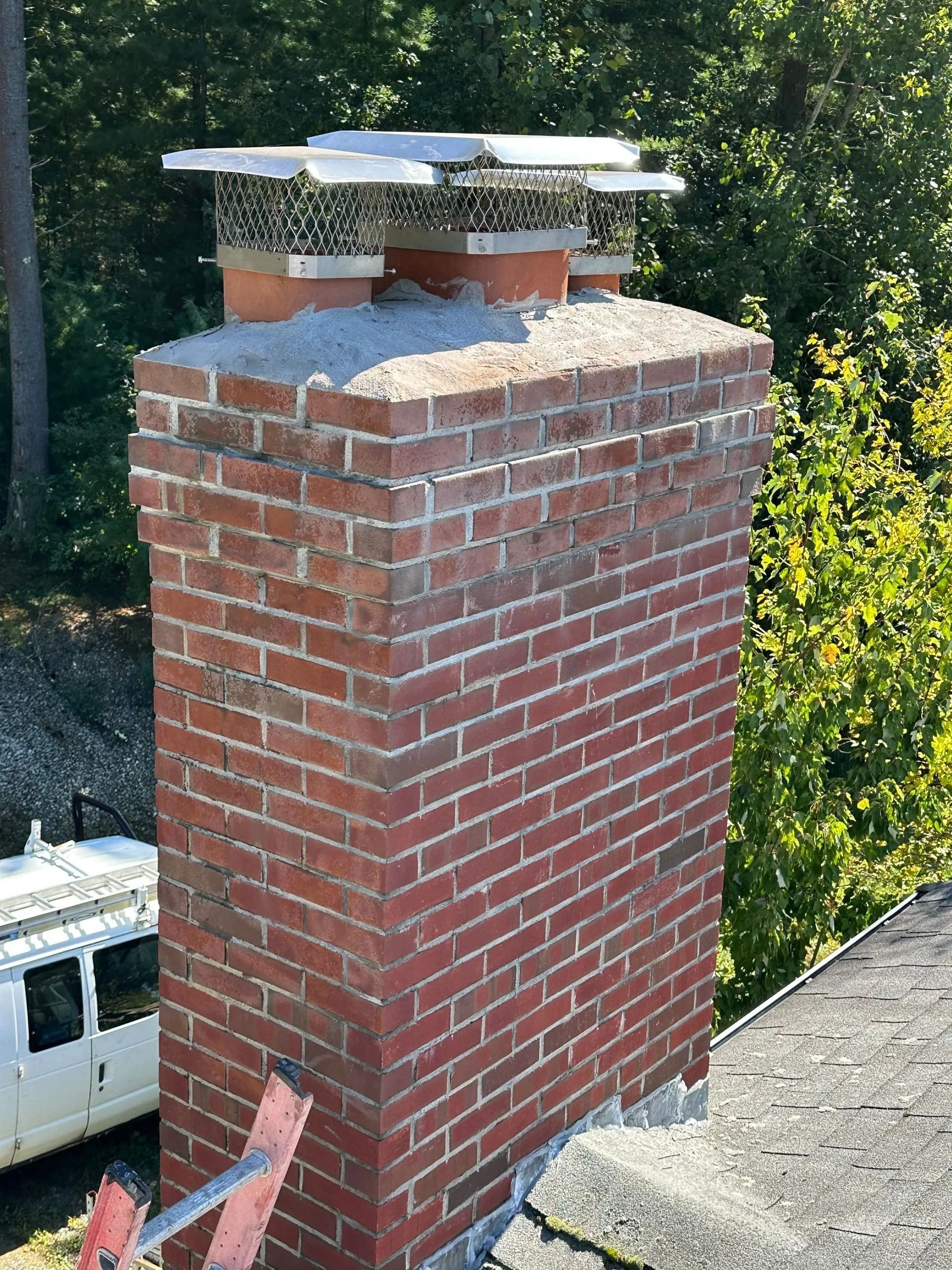 Brick chimney on a roof, topped with metal chimney caps and a concrete cap, surrounded by green trees.