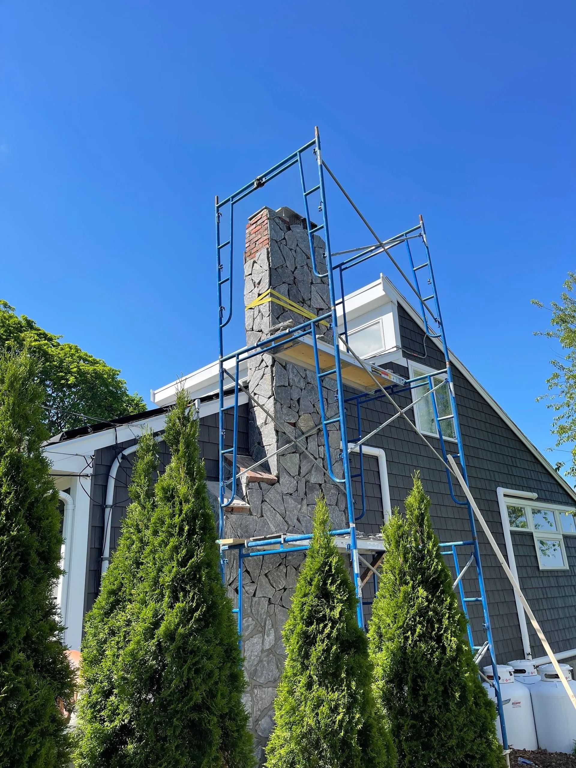 Scaffolding surrounds a brick chimney on a house with dark gray shingles under a blue sky.