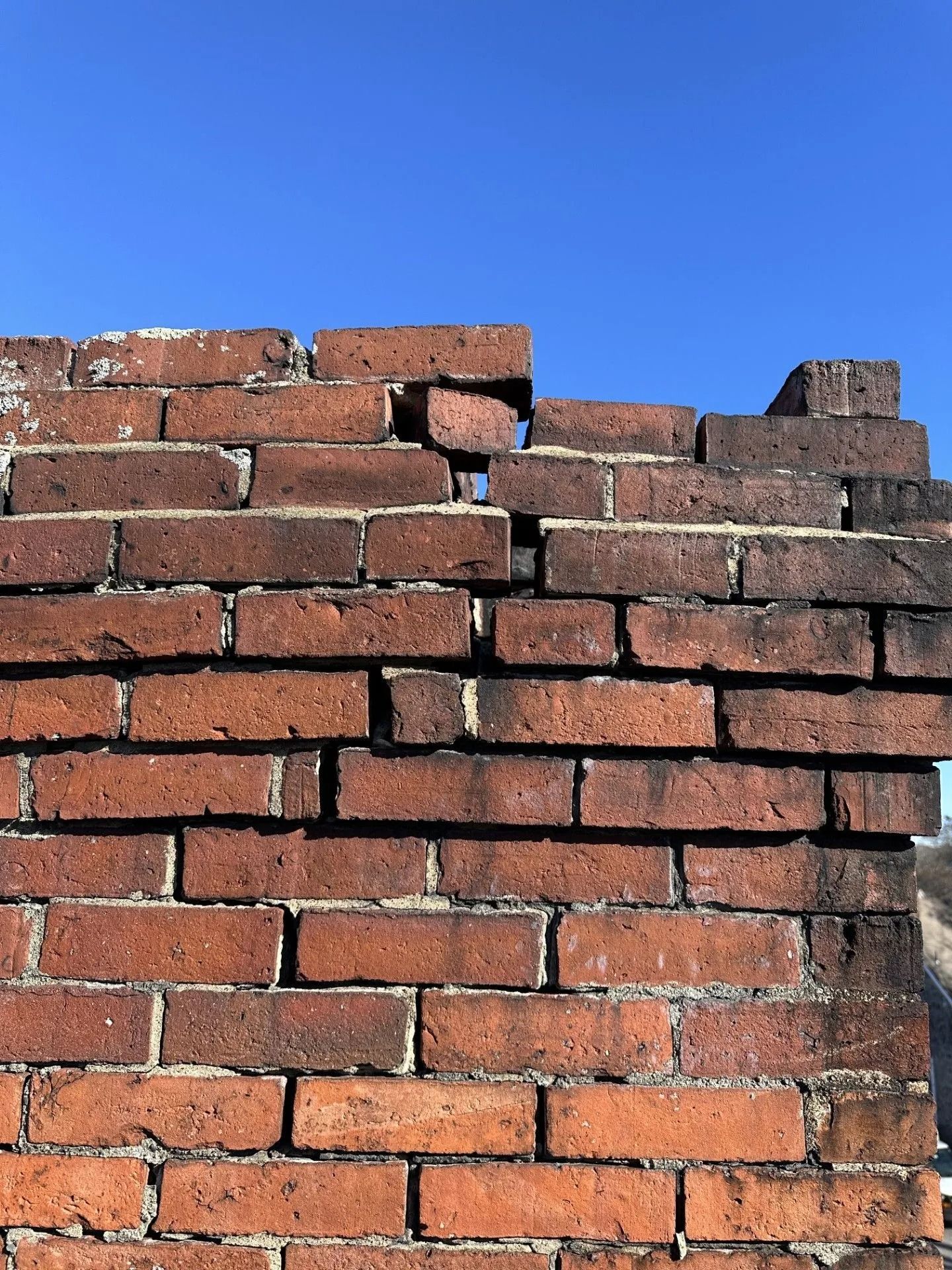 Close-up of a weathered brick chimney against a bright blue sky; some bricks are damaged or missing.