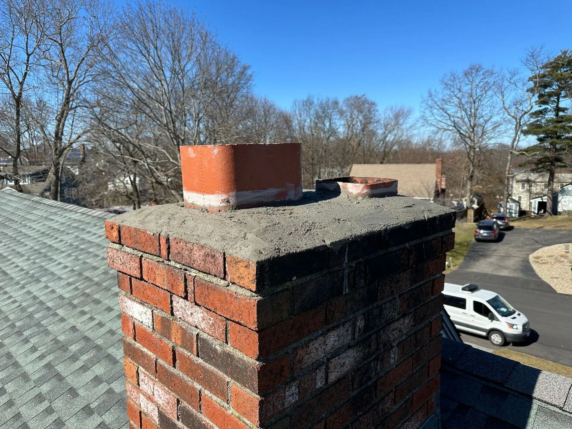 Red brick chimney on a gray shingle roof. The sky is blue. Trees and a street are in the background.