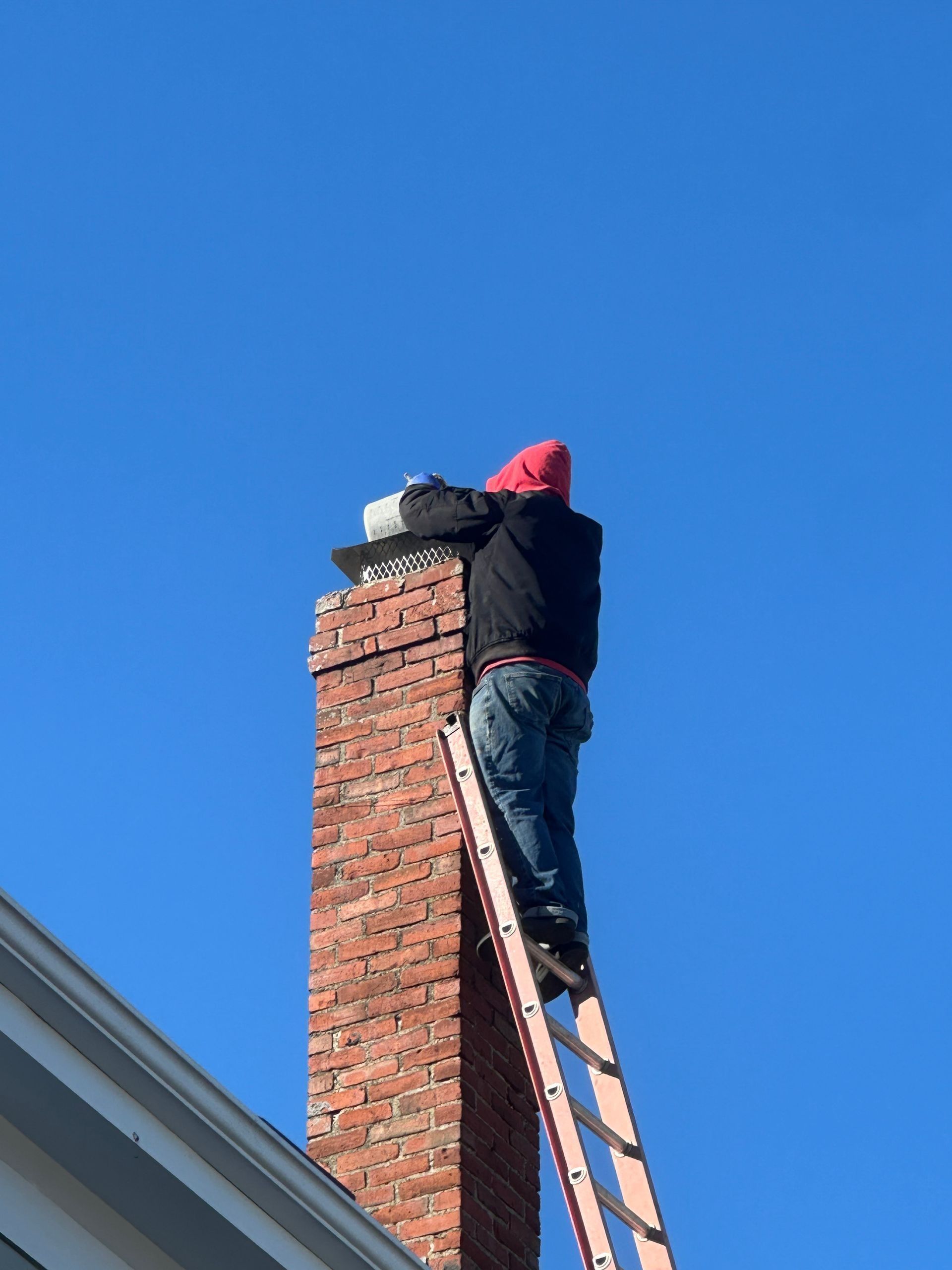 Person on a ladder, working on a brick chimney against a blue sky.