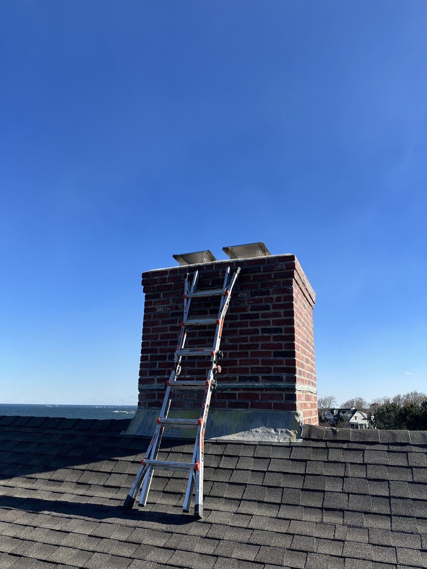 Ladder leaning against a brick chimney on a rooftop under a blue sky, with the ocean visible in the background.